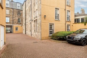 A narrow brick-paved alleyway between buildings with a parked car and a small bush.