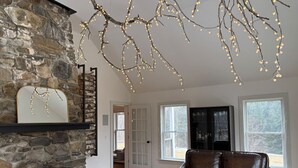 A spacious living room with a stone fireplace, leather sofa, and large windows. - Timber Tide Retreat Just 15 Minutes to Belfast (Swanville)