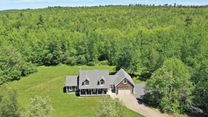 A large, two-story house with a garage, surrounded by a lush green landscape and dense forest. - Timber Tide Retreat Just 15 Minutes to Belfast (Swanville)