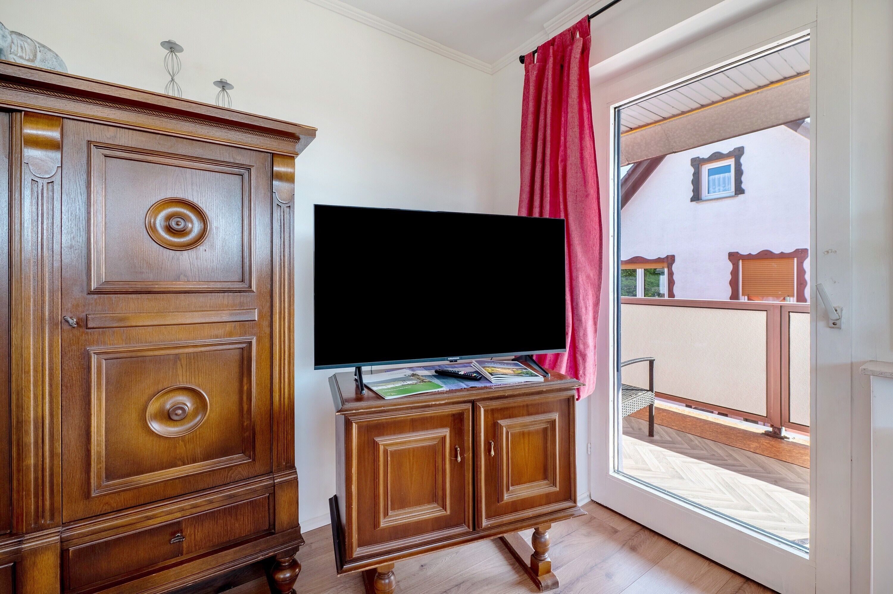 A wooden cabinet with a flat-screen TV on top, a red curtain, and a view of a balcony with a railing.