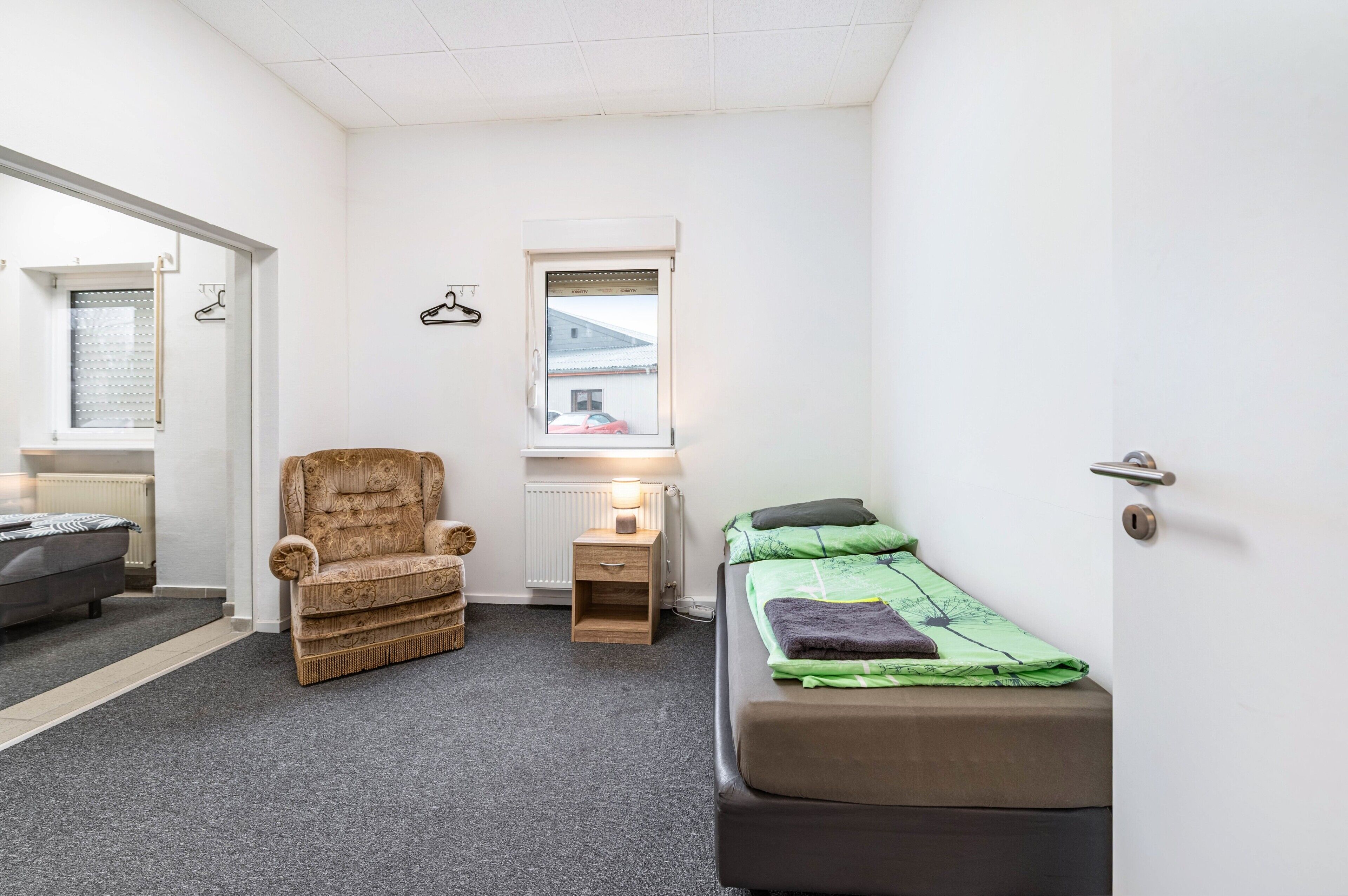 Una habitación pequeña y bien iluminada con sofá, cama, sillón, mesita de noche y ventana con vista a un coche.