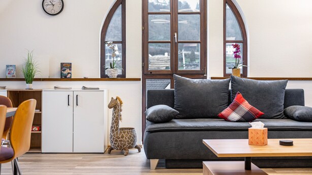 Een woonkamer met een bank, een salontafel, een boekenkast, een klok en grote ramen.