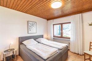 Ein Zimmer mit einem Bett, einem Nachttisch, einem Fenster mit Vorhängen und einer Holzbalkendecke.
