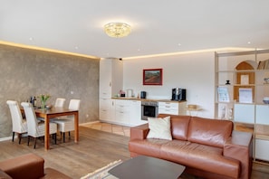 Un salon moderne avec un canapé en cuir marron, une table à manger avec des chaises à rayures, un coin cuisine avec des meubles blancs et un lustre.