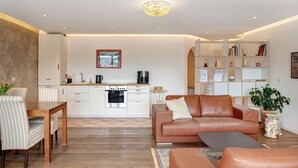 Een moderne woonkamer met een bank, een eettafel met stoelen, een keuken met witte kasten en een boekenkast.
