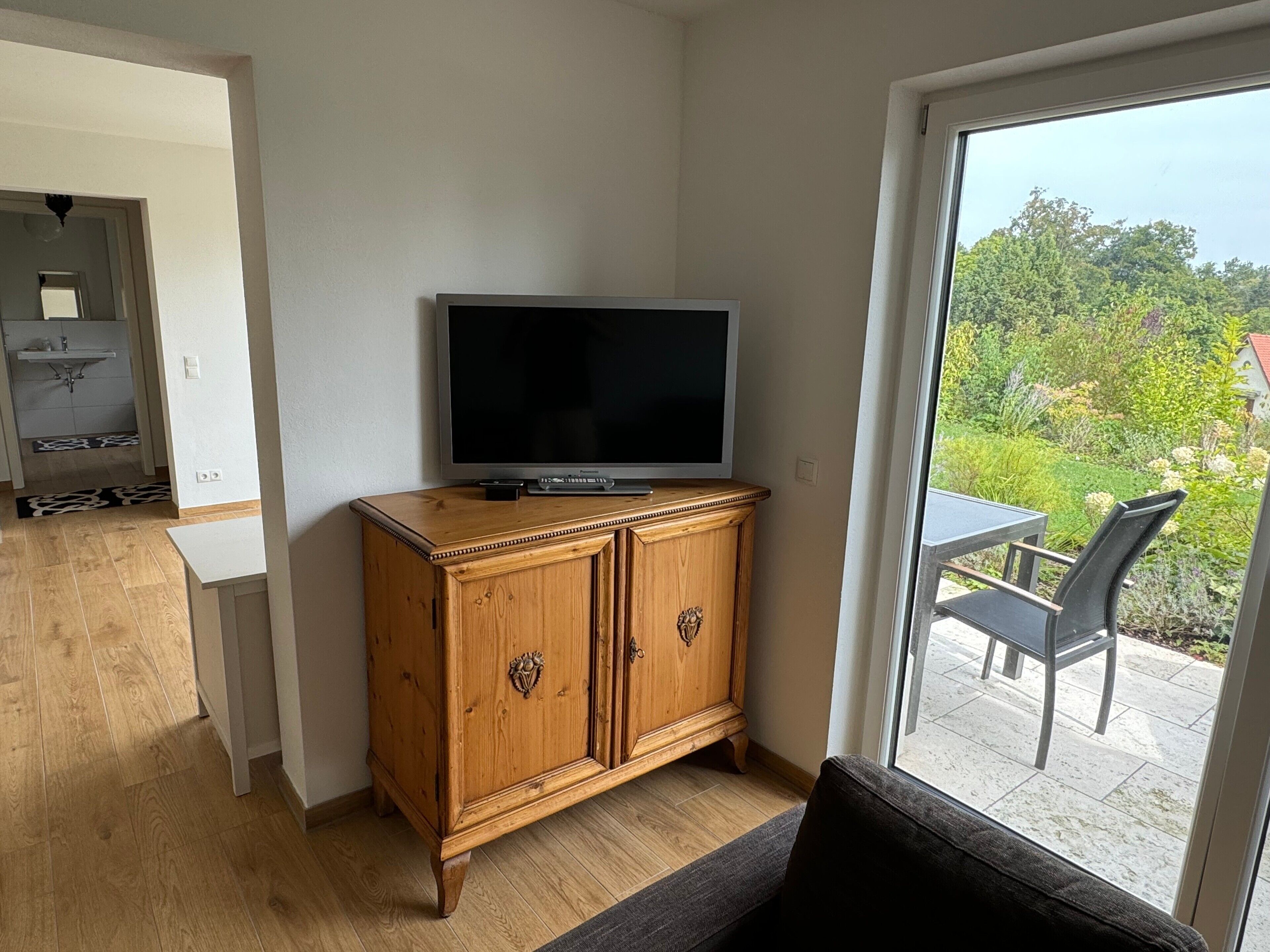 A room with a television on a wooden cabinet, a sofa, and a view of a garden through a glass door.