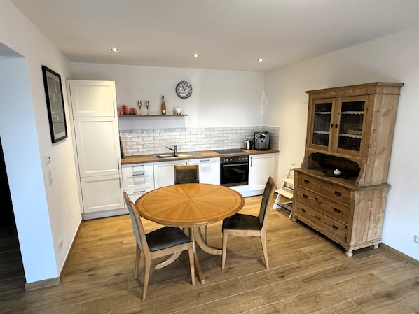 Una cocina compacta con una mesa redonda de madera, armarios blancos, una cafetera y un reloj en la pared.