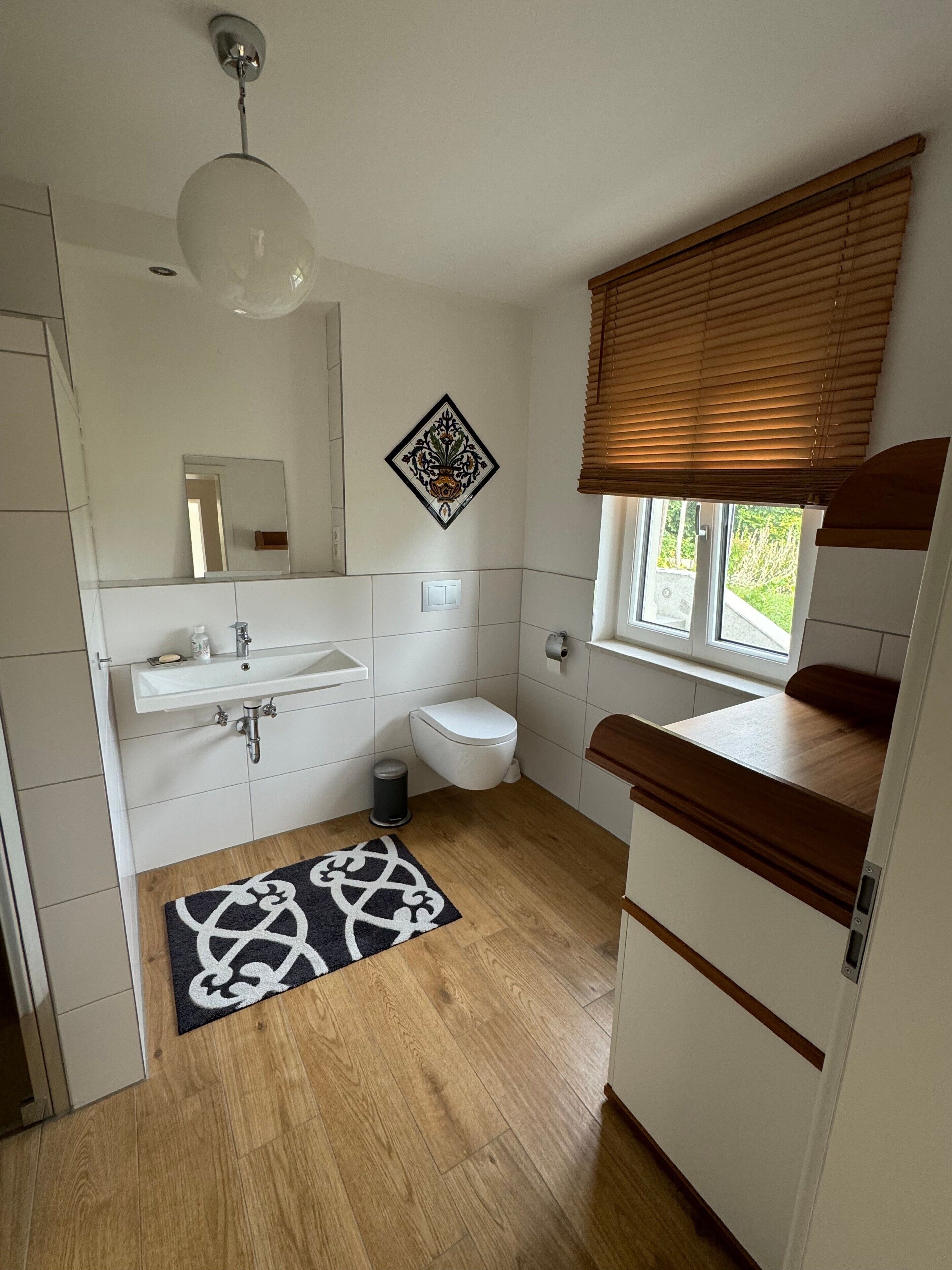 Un baño con lavamanos, inodoro y tocador blancos, suelo de madera, alfombra blanco y negro, y una ventana con persianas.