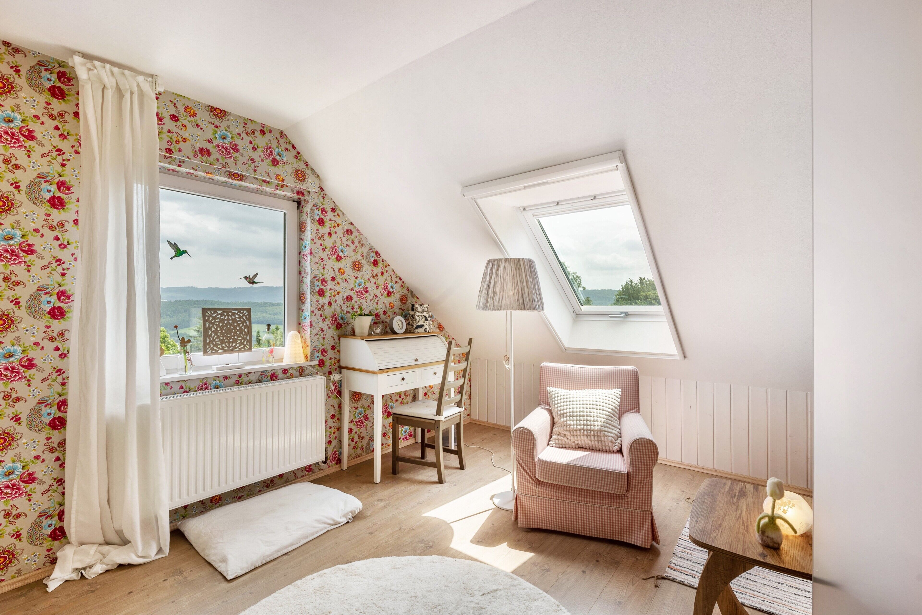 Una habitación acogedora con techo inclinado, un escritorio, una silla, un sillón con cojín, un radiador y una ventana con vistas.