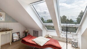 Ett rum med ett sluttande tak, en säng med ett rött täcke, ett skrivbord med en nallebjörn, en inramad tavla och ett fönster med utsikt över träd.