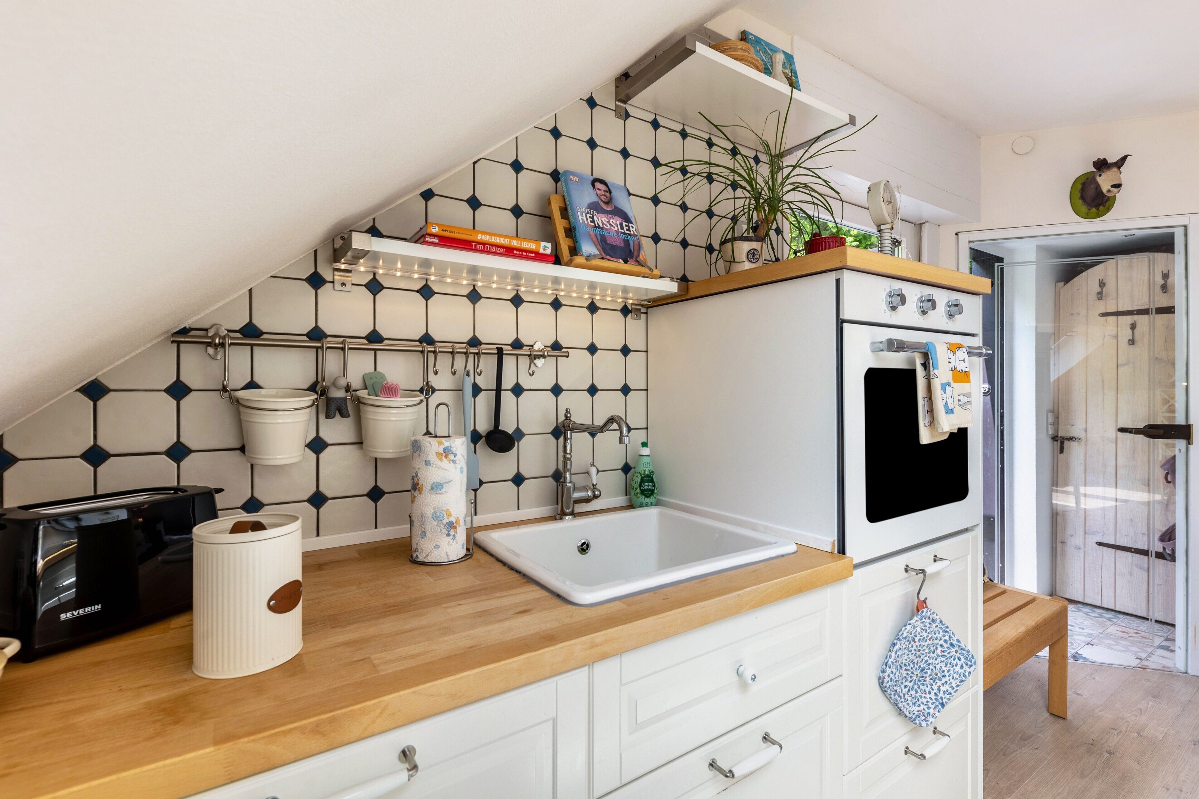 Une cuisine avec un plan de travail en bois, des placards blancs, un évier blanc et une cuisinière. On peut voir des livres, une plante et des ustensiles de cuisine sur les étagères.