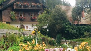Une maison traditionnelle en bois, dotée d’un balcon et entourée d’un jardin où poussent différentes variétés de fleurs.