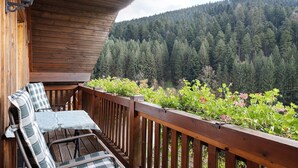 A wooden balcony with a view of a forested hillside, a table with chairs, and a railing.