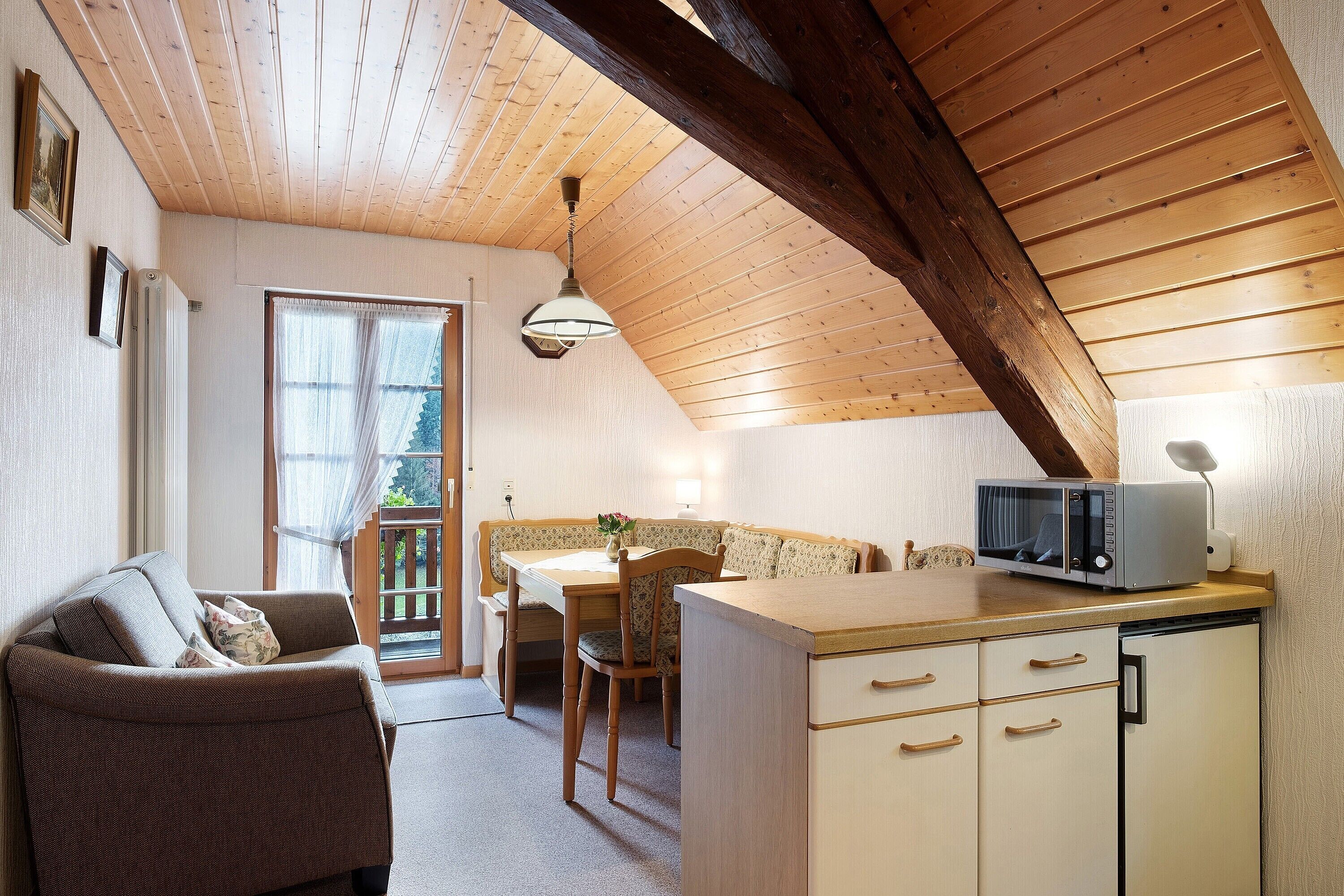 A cozy room with a wooden ceiling, a dining table with chairs, a sofa, and a kitchenette with a microwave.