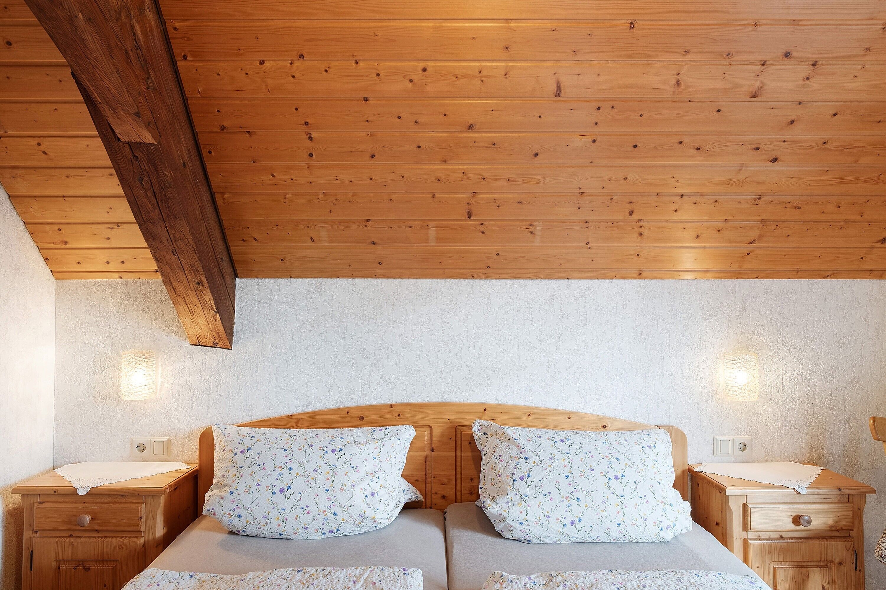 A room with a wooden ceiling, two beds with floral bedding, bedside tables, and wall-mounted lights.