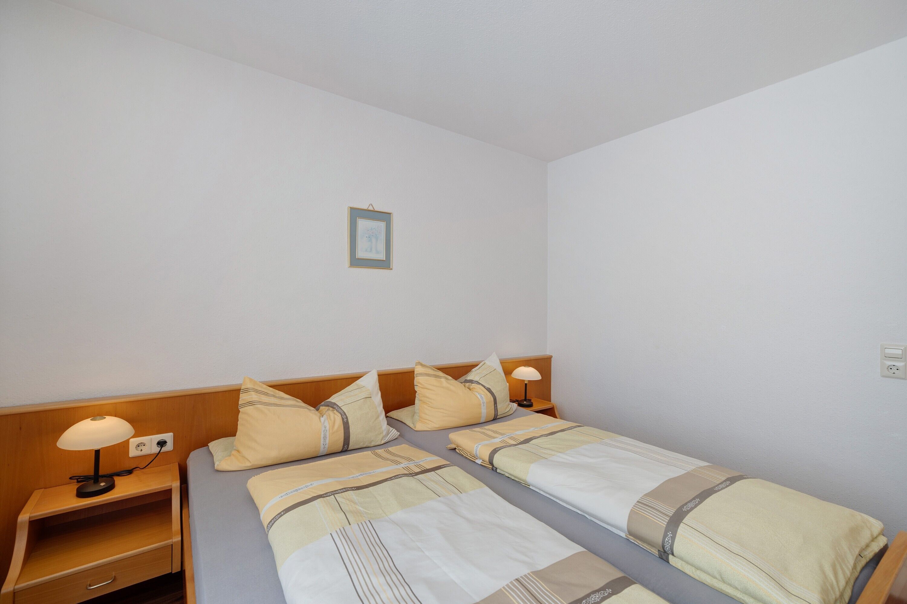 Habitació d'hotel amb dos llits, una tauleta de nit amb una làmpada, un quadre amb marc a la paret i una presa elèctrica.