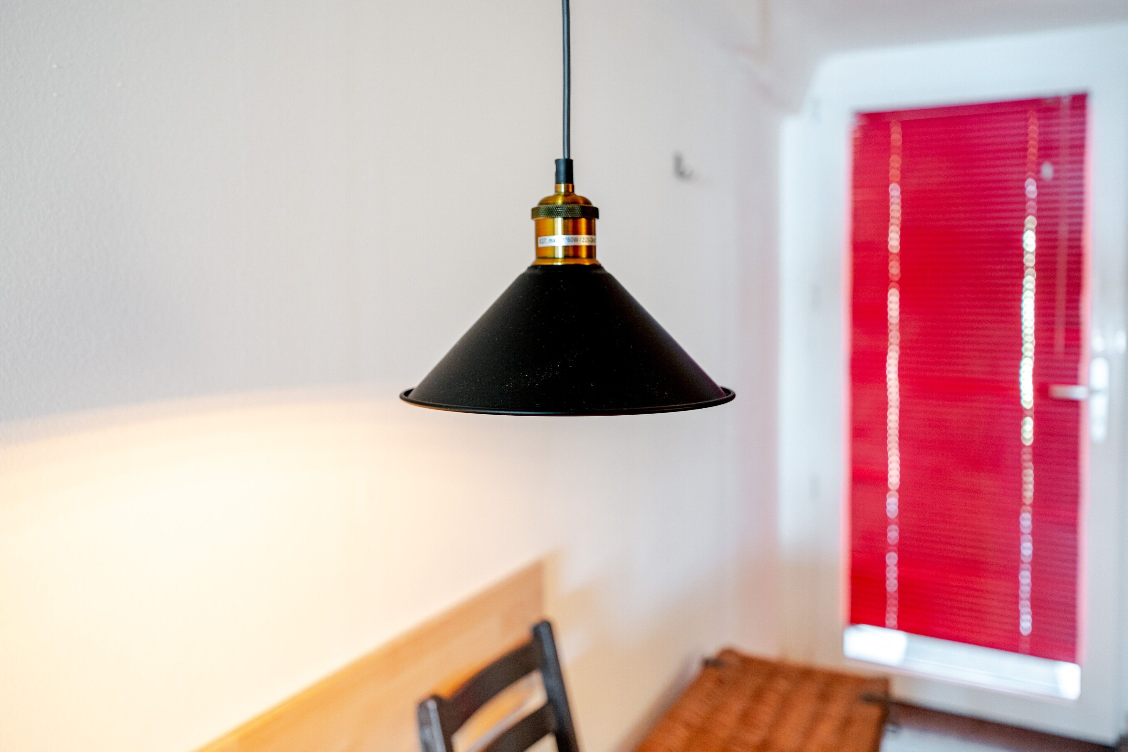 A black pendant light hanging from the ceiling in a room with a wooden chair and a window with red blinds.