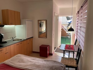 Een kleine keuken met een spoelbak, een kleine koelkast, houten kastjes, een rode kast, een spiegel, en een eethoek met een tafel en stoel.