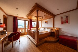 A four-poster bed with a wooden headboard, a desk with a chair, a red bench, and a view of the outdoors through a window.