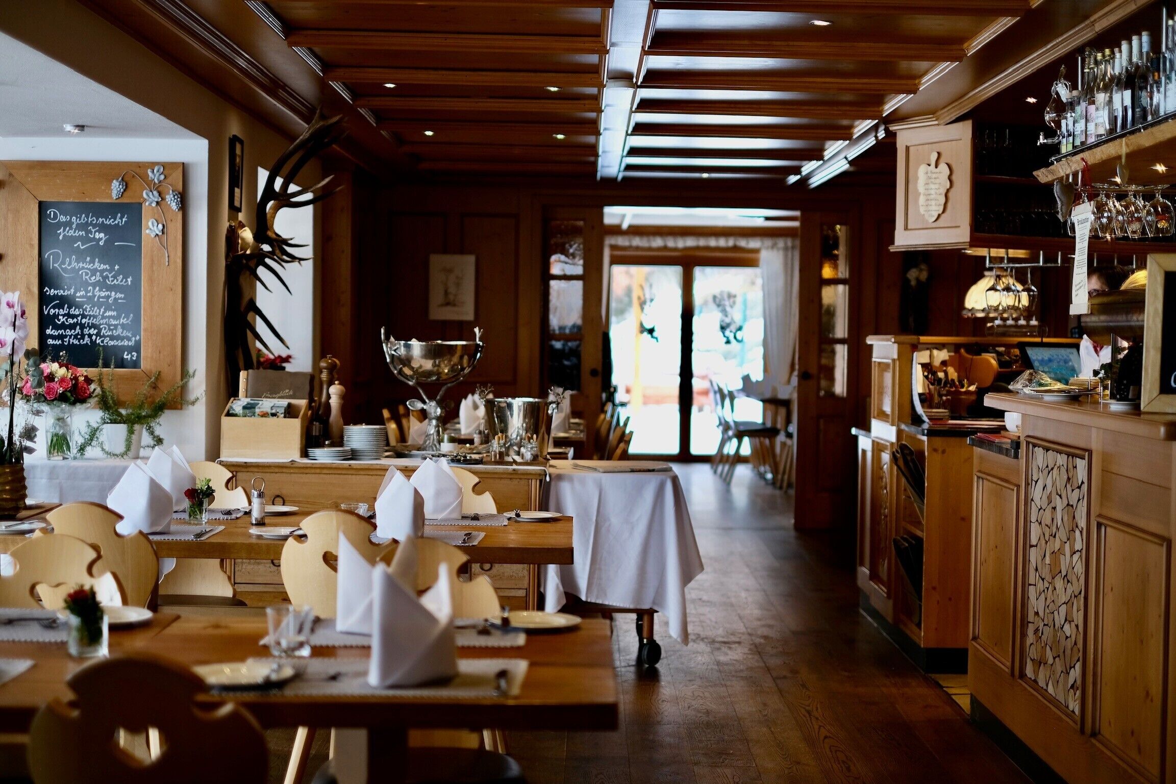 A restaurant with wooden tables and chairs, a bar area with bottles and glasses, and a chalkboard menu on the wall.