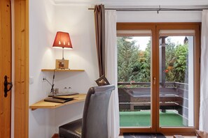 A room with a wooden desk, a chair, a lamp, and a sliding glass door leading to an outdoor area with a green lawn and wooden deck.