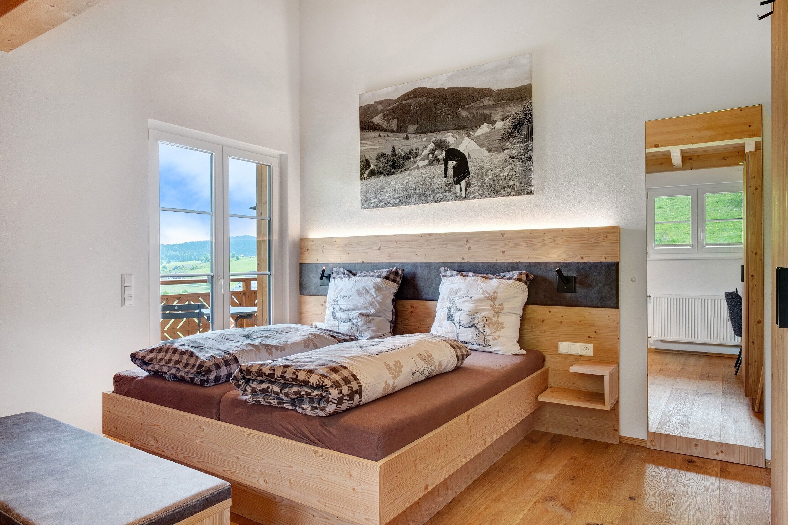 Una camera da letto con un letto in legno, vista sull'esterno, un quadro appeso al muro e una piccola mensola in legno.