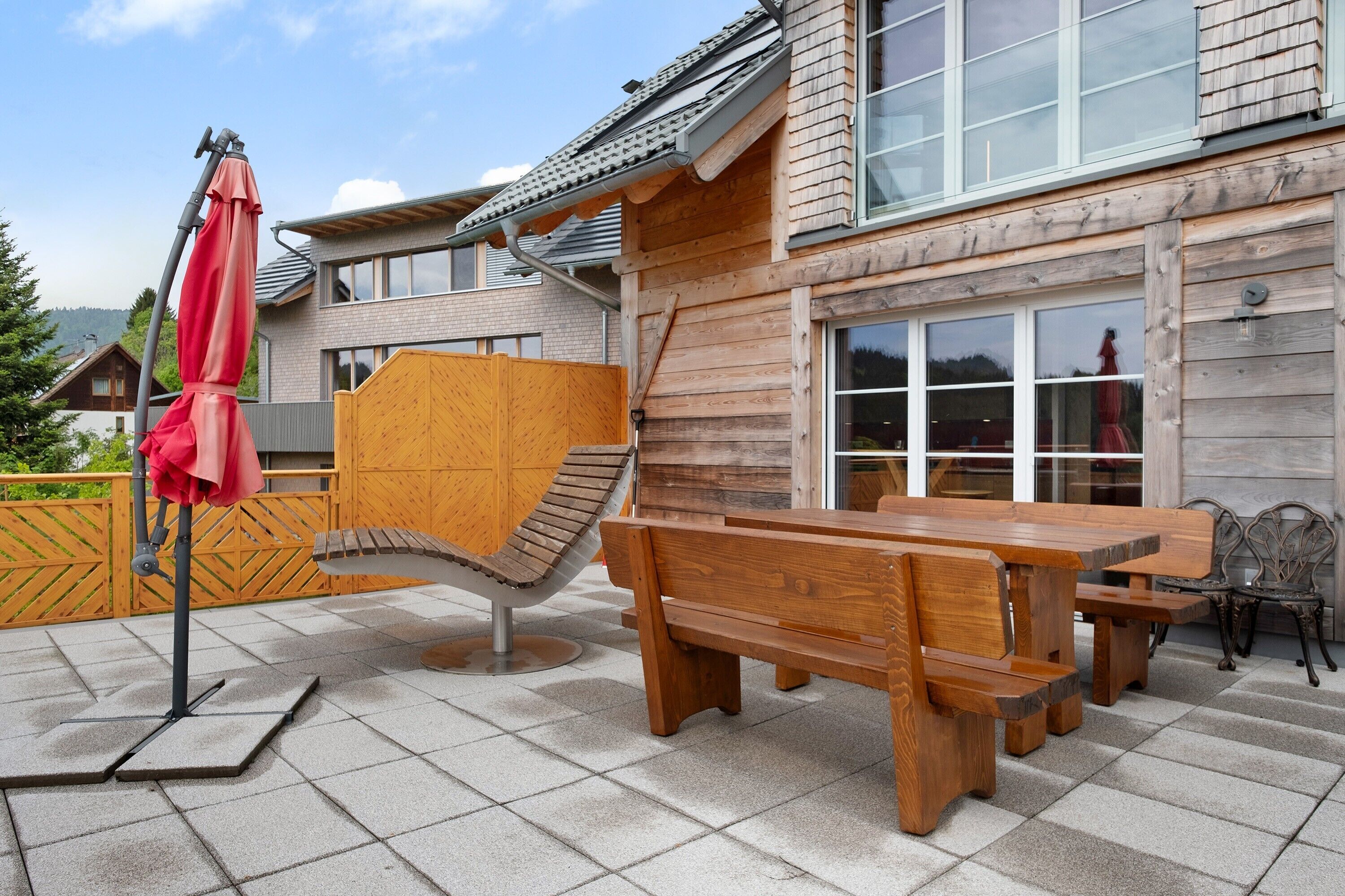Un terrazzo in legno con una panca e un tavolo, un ombrellone rosso, e una casa con tetto spiovente.