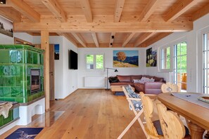 Un techo con vigas de madera, una chimenea con azulejos verdes, un sofá de cuero marrón y una mesa de comedor de madera con sillas.