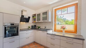 Une cuisine moderne dotée d’un four encastré, d’une fenêtre avec stores et offrant une vue sur la verdure environnante.