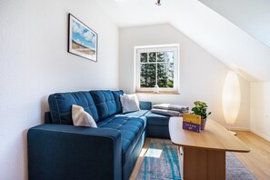 Un salon moderne avec un canapé bleu, une petite table sur laquelle est posée une plante, un tableau encadré au mur et une fenêtre donnant sur des arbres.