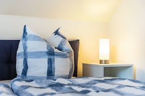 Un lit recouvert d’une couverture à carreaux bleus et blancs, un oreiller et une table de chevet avec une lampe.
