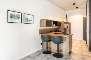 A modern kitchen with a wooden island and bar stools, framed artwork on the wall, and a tiled floor.