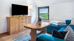Ett rum med en tv-apparat på ett träskåp, ett bord med en lampa, ett fönster med utsikt och en blå soffa med kuddar.