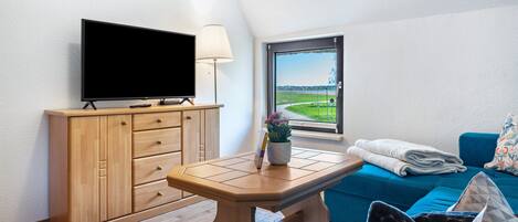 A room with a television on a wooden cabinet, a table with a lamp, a window with a view, and a blue sofa with pillows.