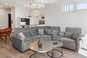 Ein modernes Wohnzimmer mit einem grauen Ecksofa, einem runden Couchtisch und einem Essbereich mit Holztisch und orangefarbenen Stühlen.