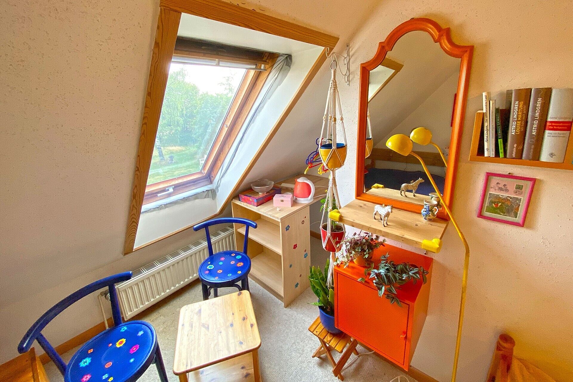 Een kinderkamer met een schuine kap, een raam, een spiegel, een bureau, een stoel en een boekenplank.