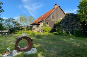 Una casa con techo inclinado, una gran estructura de madera a la derecha y una escultura metálica circular en primer plano.