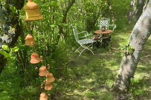 Un jardín con una mesa y silla de metal, rodeado de follaje y campanillas colgantes.