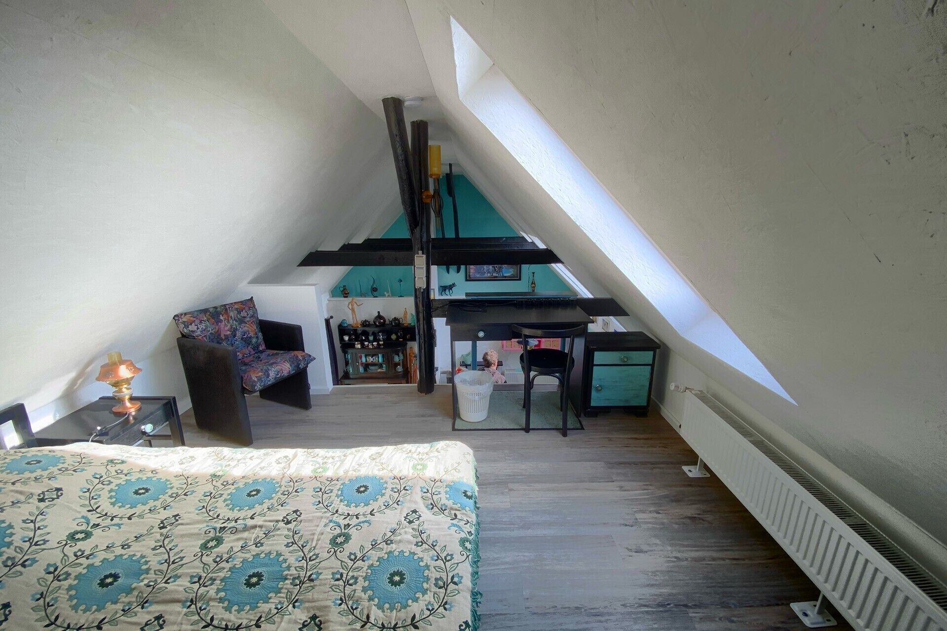 A room with a sloped ceiling, a bed with a patterned bedspread, a chair, a table, and a shelf with decorative items.