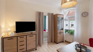 A room with a TV, wooden cabinet, dining table, red chair, and a window with curtains.