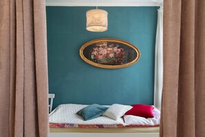 A bedroom with teal walls, a bed with white and patterned bedding, a framed floral artwork, and a hanging light fixture.
