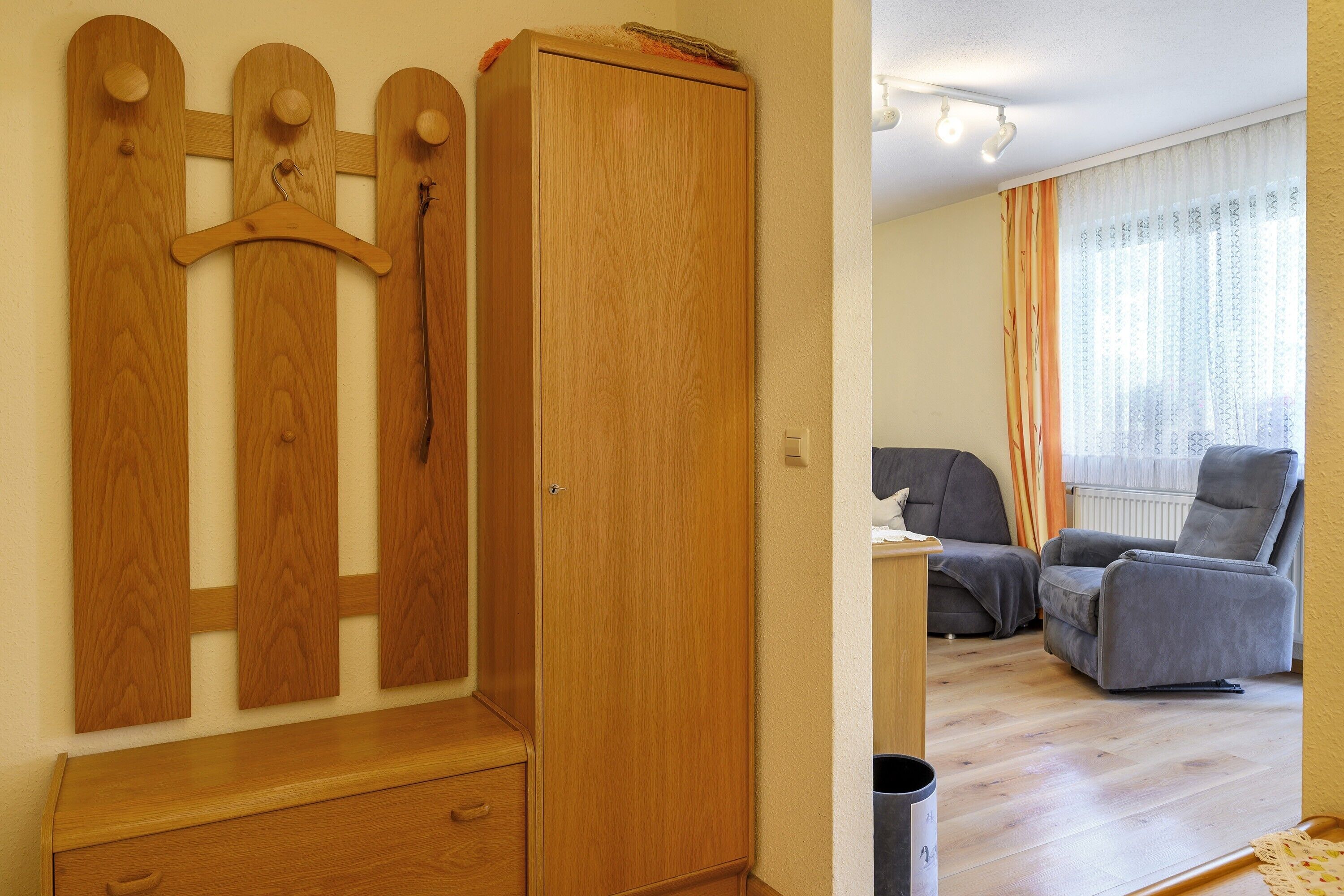 Une pièce comprenant une armoire en bois, un banc en bois, un fauteuil gris et une poubelle noire.