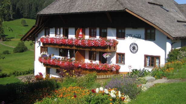 Ein traditionelles Gebäude im alpinen Stil mit einem Balkon, der mit Blumen, einer Uhr und einem Schild mit dem Namen und dem Gründungsjahr des Hauses geschmückt ist.