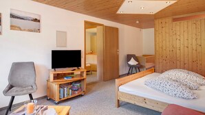 Ein Zimmer mit Holzboden, einem Bett mit floralem Bettbezug, einem Fernseher auf einem Holzständer, einem Sessel und einem Bücherregal.