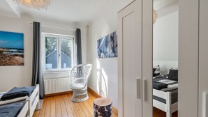 Un dormitorio con cama, una silla, un taburete de madera redondo, un radiador y una ventana con cortinas.