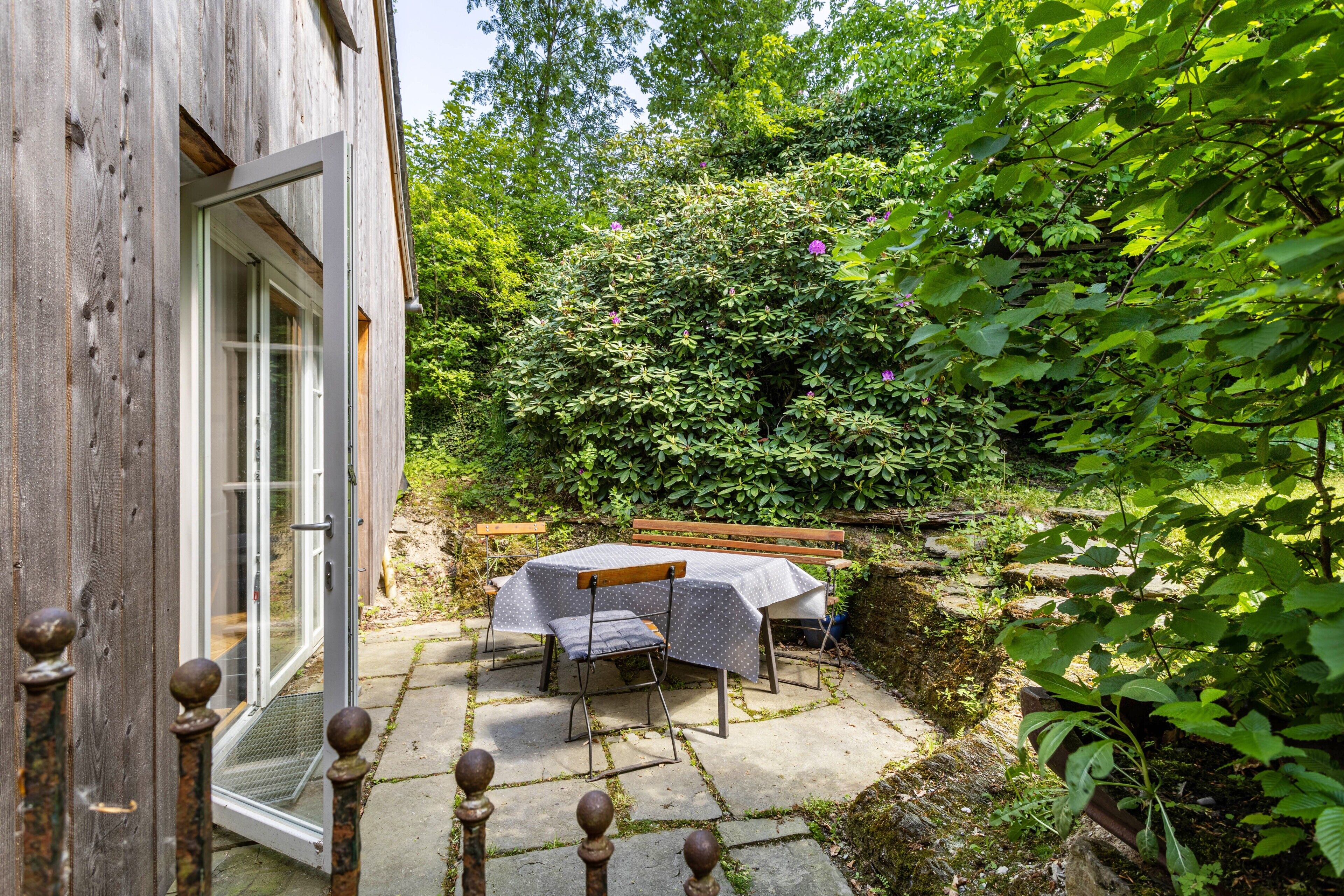 A wooden building with a glass door, a stone patio with a table and chairs, and a bench.