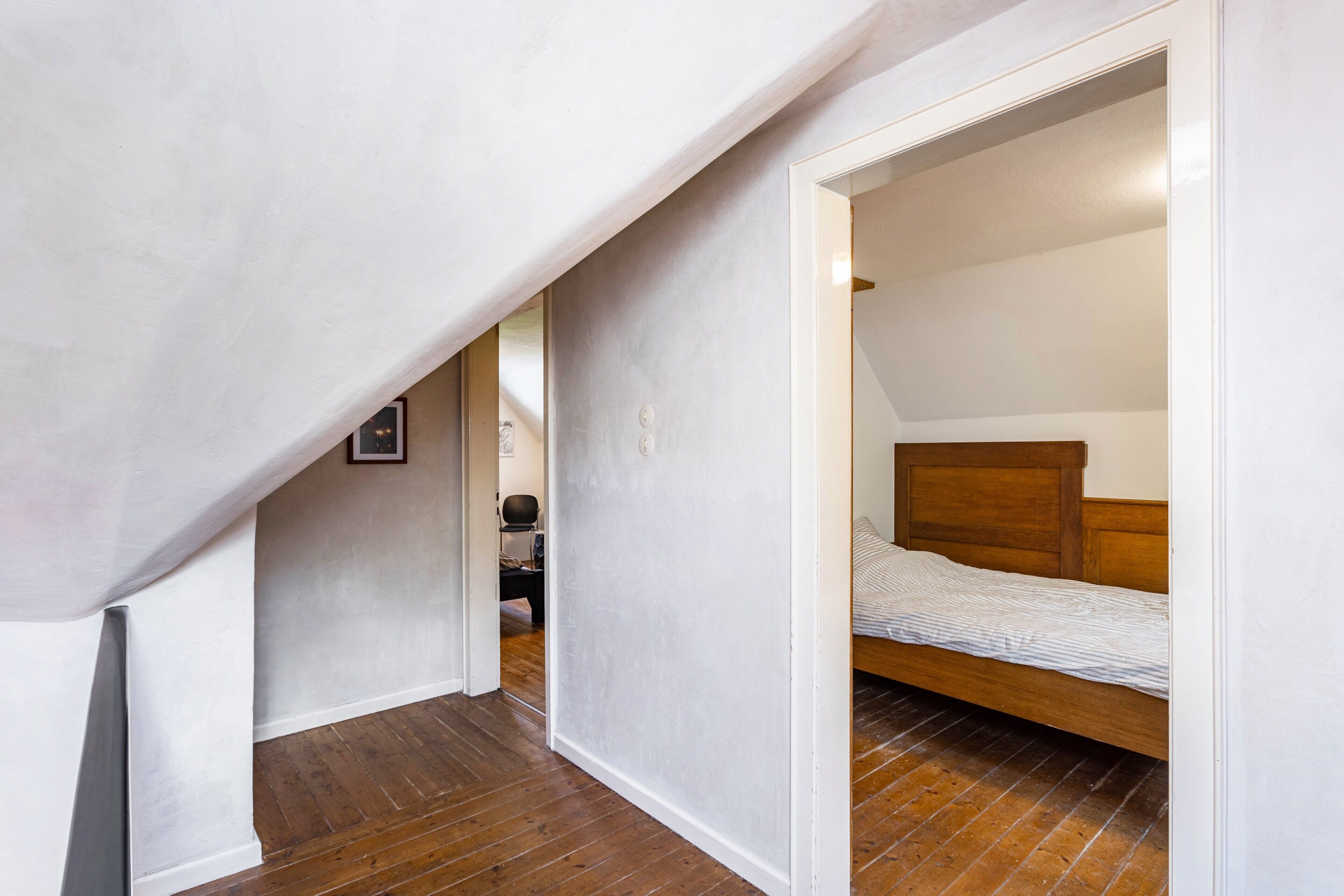 Un estrecho pasillo con piso de madera que conduce a un dormitorio con estructura de cama de madera y colchón blanco.