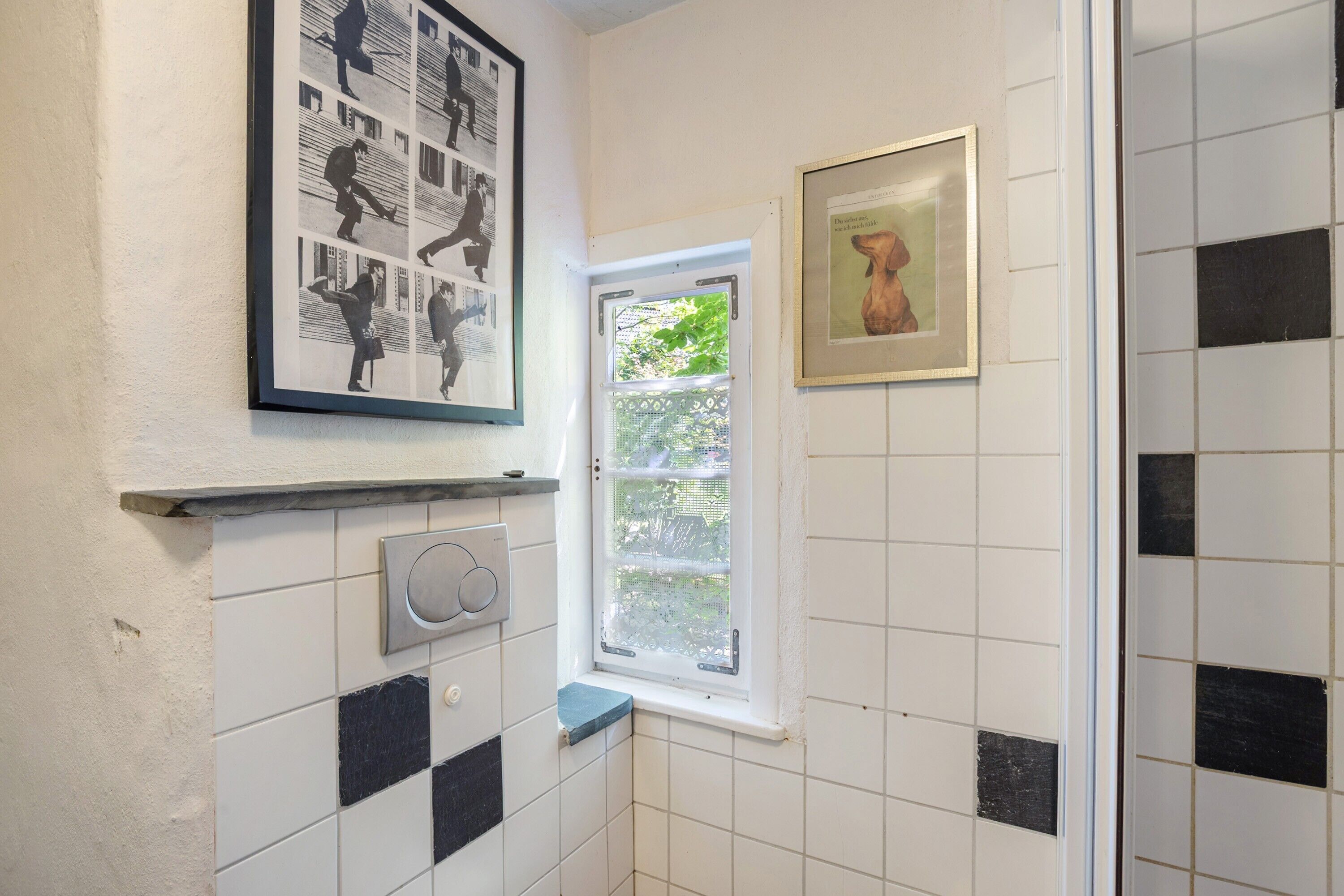 Une salle de bain avec des carreaux noirs et blancs, une fenêtre, un tableau encadré et un interrupteur fixé au mur.