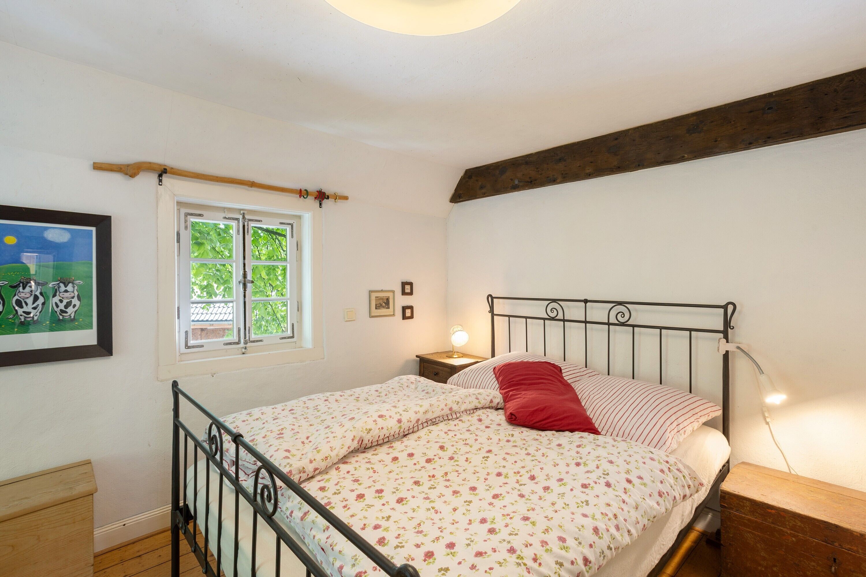 Ein Schlafzimmer mit Bett, Fenster, Nachttisch, Lampe und einem Bild an der Wand.