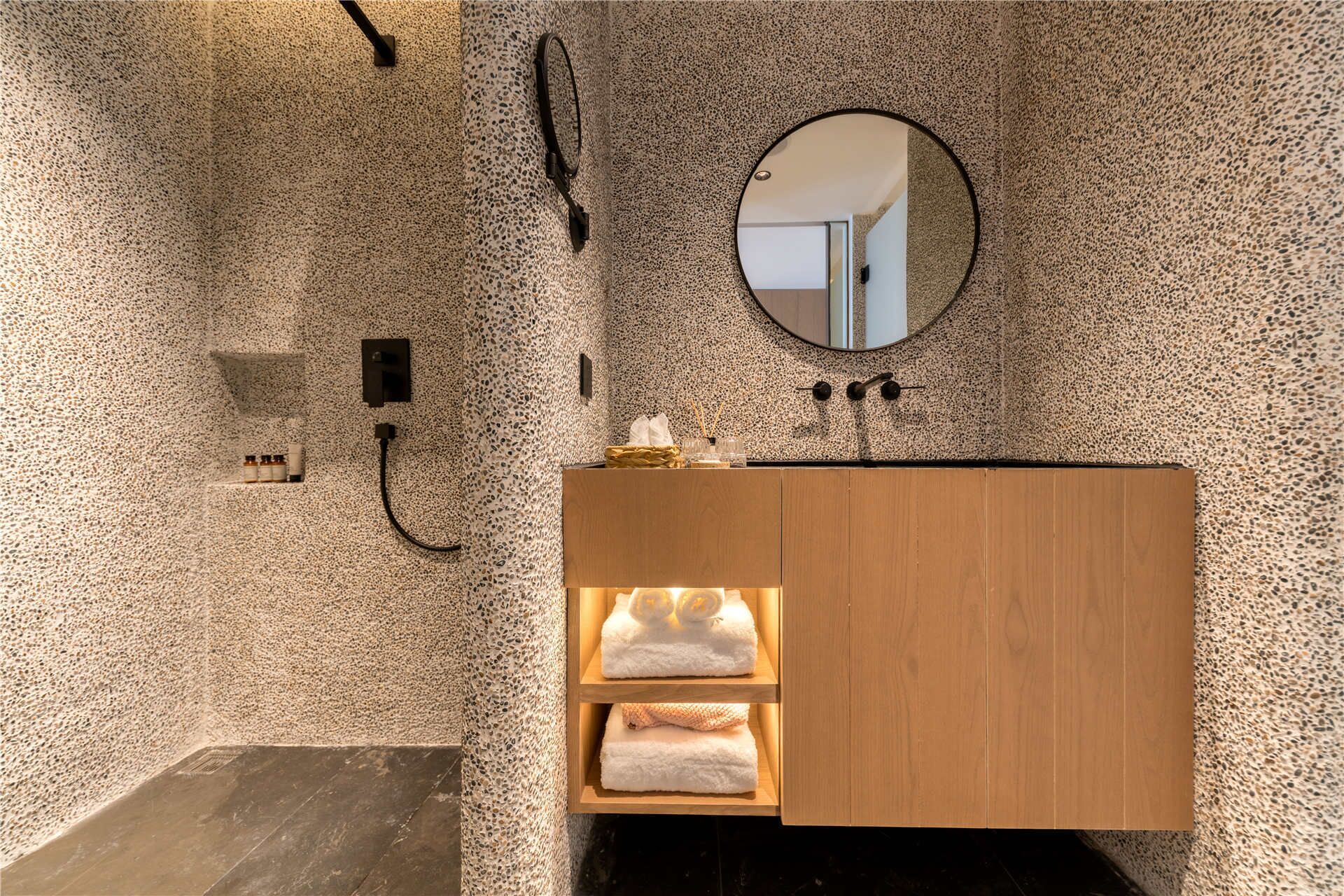 A bathroom with a speckled stone wall, a round mirror, a wooden vanity with shelves, and a black floor.